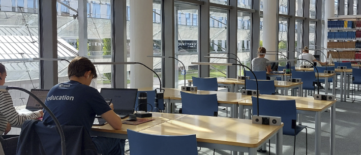 Bright library study area with large windows, individual desks, and several people working on laptops. Bookshelves line the back wall in a spacious, sunlit room.